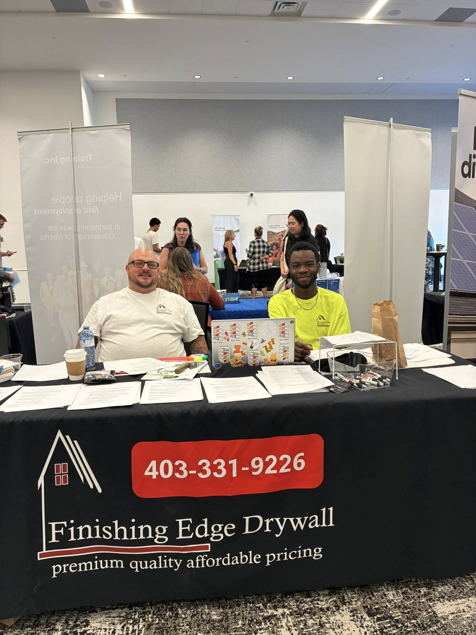 Two men sit at a booth with "Finishing Edge Drywall" branding at a busy expo. Papers, a laptop, and promotional items are on the table. The atmosphere is lively.
