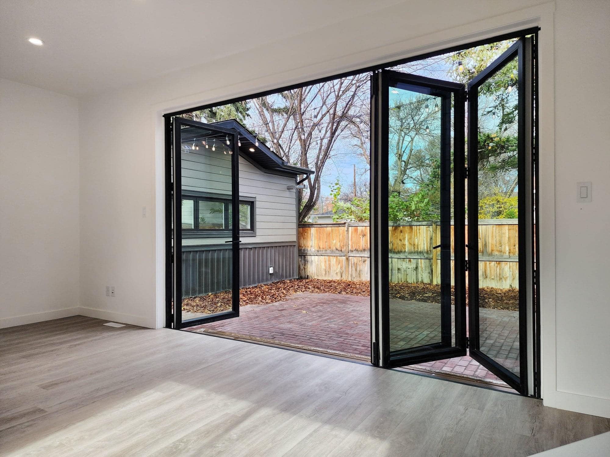 Modern room with large folding glass doors opening to a patio with wooden fencing and greenery. Bright, inviting space with light wood floors.