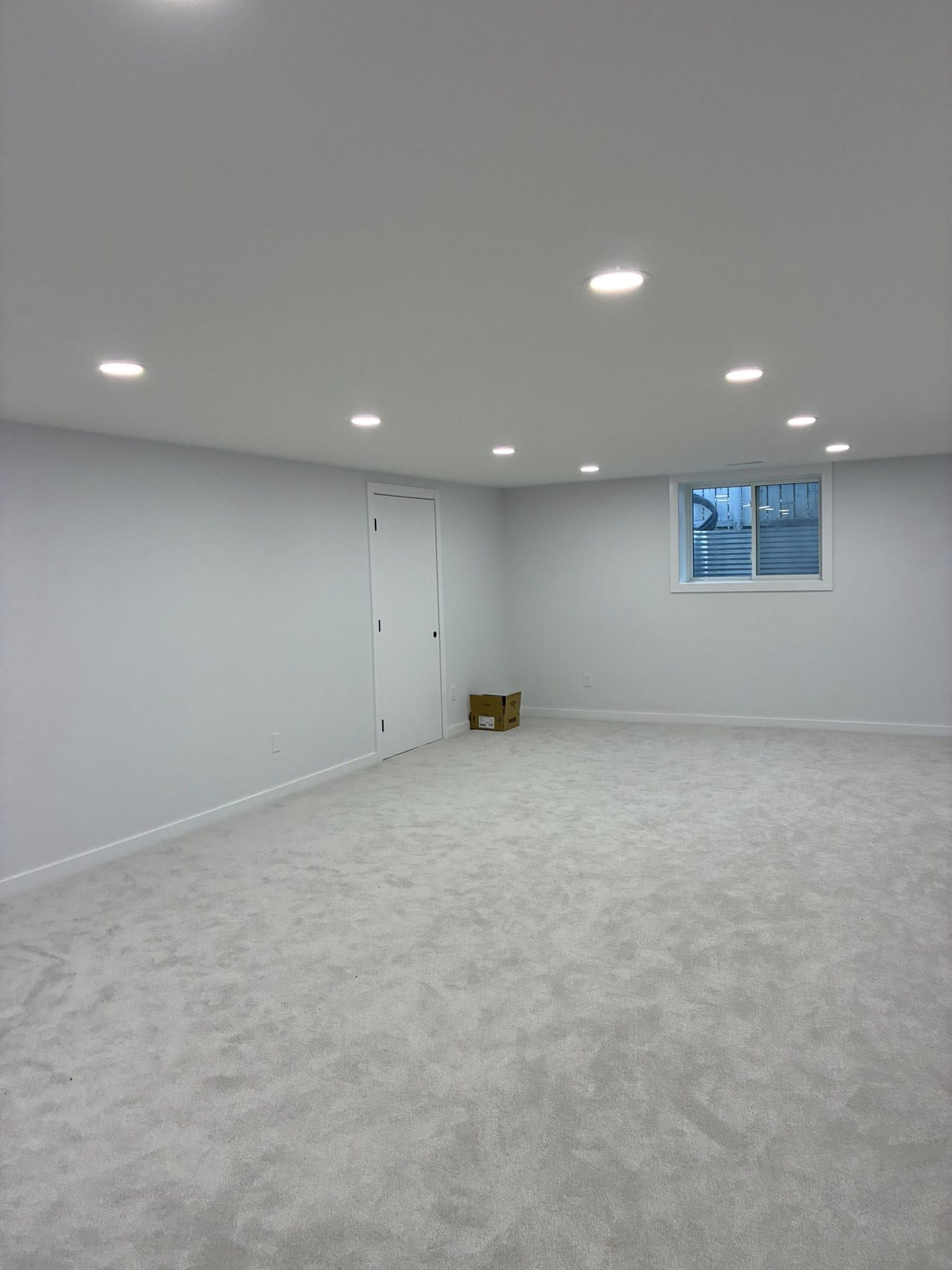 Spacious, minimalistic room with white walls, carpeted floor, and recessed ceiling lights. A single window and a cardboard box sit against the far wall.