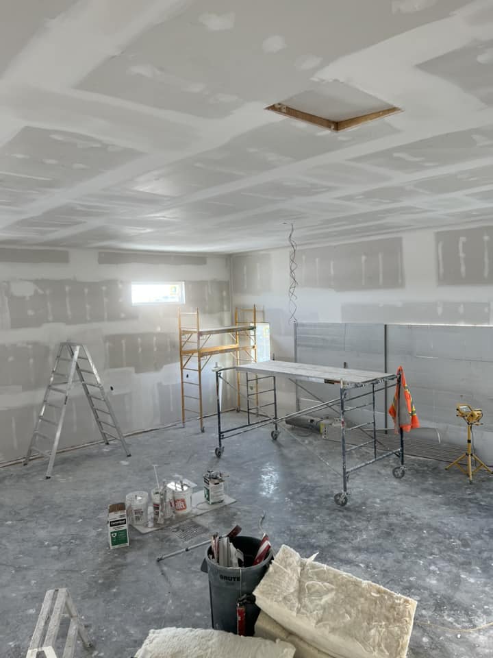 Construction site with unfinished drywall and scaffolding. Items include a ladder, buckets, and tools on a cement floor, under bright natural light.